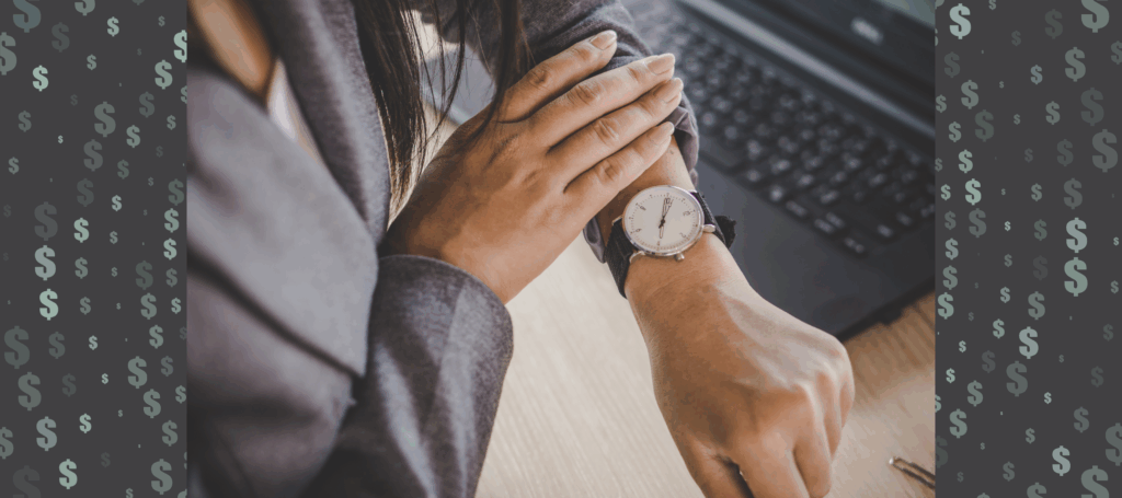 A close up of a businesswoman's arm holding up her watch as she stands at her desk in front of her laptop.