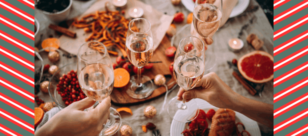 Four champagne glasses are held out, ready for a toast amidst a background of a festive table laden with food and holiday decor.